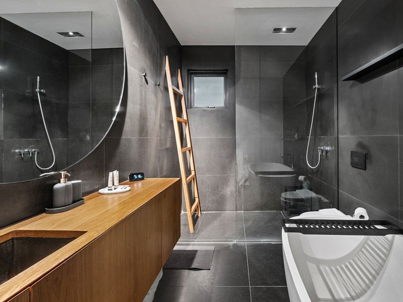 A modern and elegant bathroom featuring dark grey tiles, a wooden vanity with a large round mirror, a ladder-style towel rack, and a sleek bathtub. 
