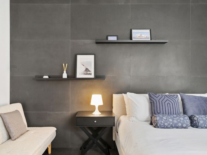 A modern bedroom with a minimalist design, featuring a bed with white and patterned pillows, a side table with a lamp, and two floating shelves on a dark gray tiled wall. 