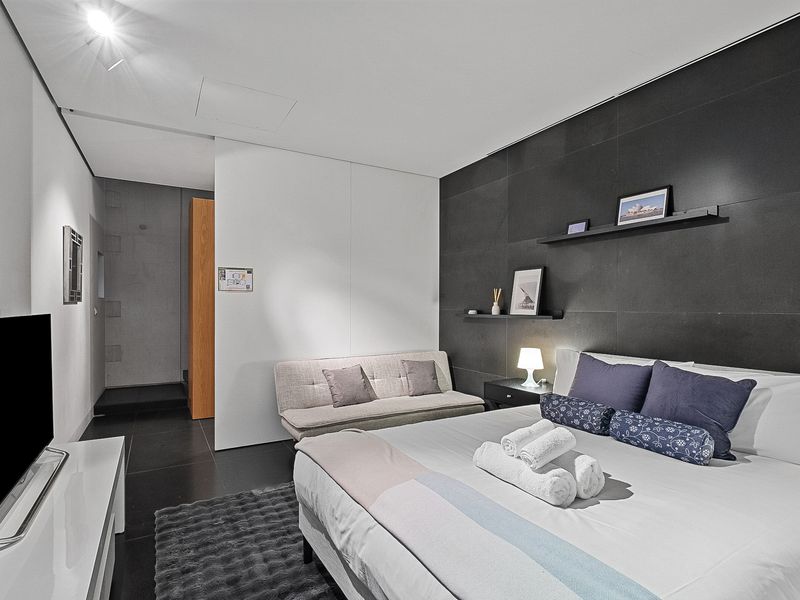 A stylish and contemporary bedroom featuring a comfortable bed with crisp white linens and accent pillows, complemented by a dark feature wall with floating shelves displaying decorative items. The room also includes a sleek flat-screen TV and a cozy sofa