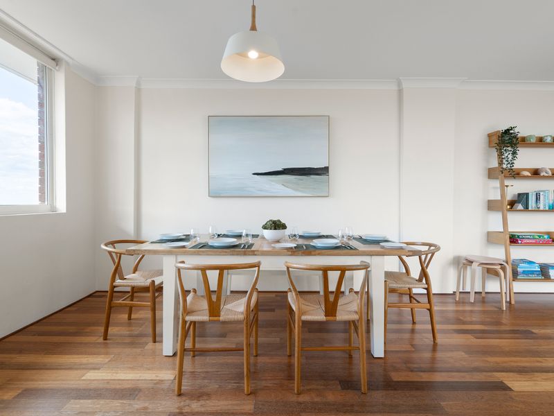 A bright, modern dining room featuring a wooden table with white chairs, set against a white wall with minimalist artwork and large windows.