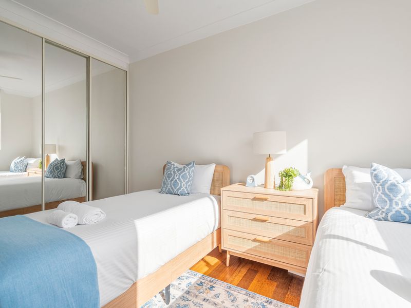 A brightly lit bedroom with two single twin beds separated by a light wood three-drawer nightstand.