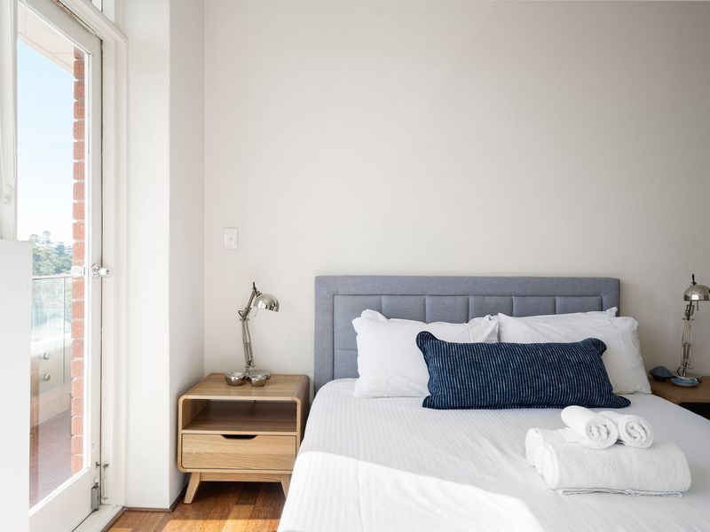 An airy, modern bedroom with a neatly made bed, natural light from a glass balcony door, and clean white walls.