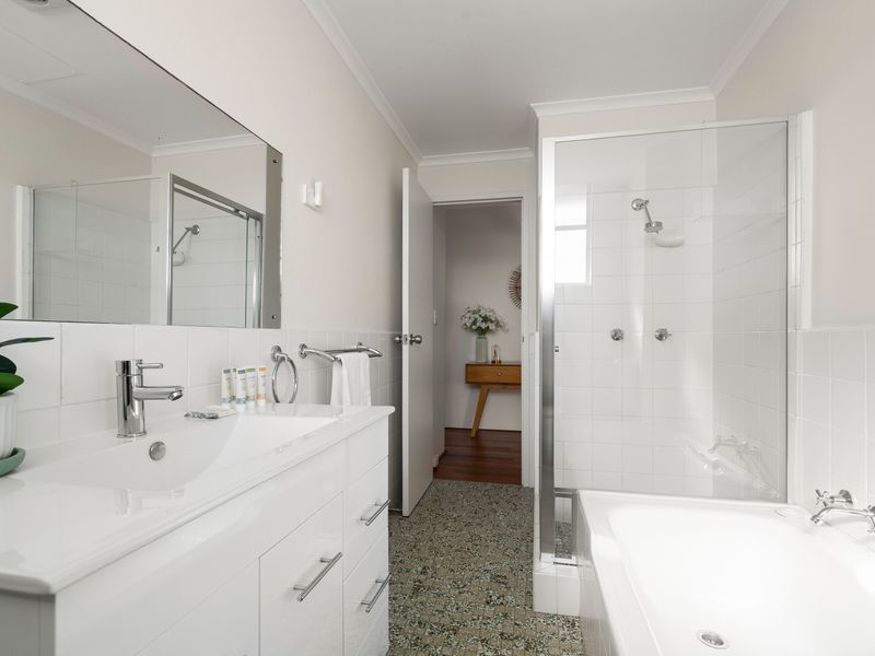 A modern, all-white bathroom featuring a vanity with a large mirror, a separate bathtub, a glass walk-in shower, and speckled floor tiles.
