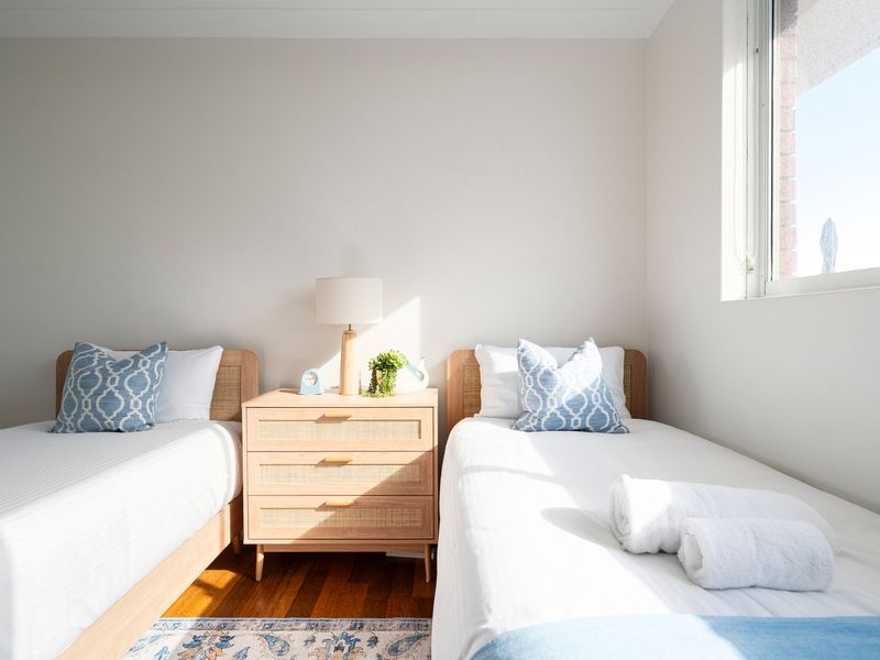 Bright, sun-drenched twin bedroom featuring crisp white linens, serene coastal blue accents, and a modern light-wood dresser.
