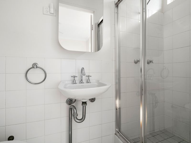 A modern, compact white tiled bathroom featuring a wall-mounted sink with exposed chrome plumbing, a frameless mirror, and a glass-enclosed shower.