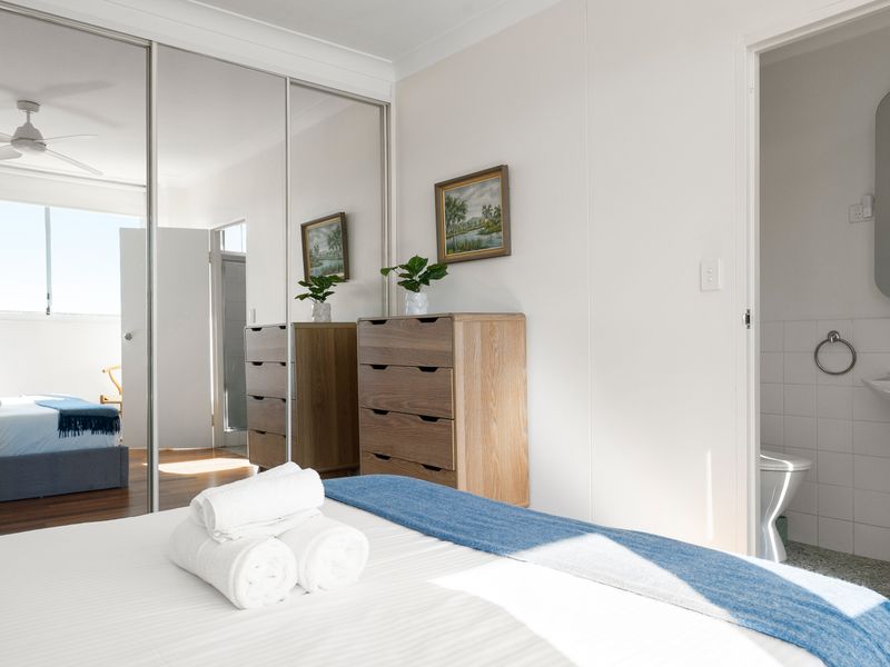 Bright bedroom with white linens, a blue throw, and a mirrored wardrobe next to an ensuite bathroom.