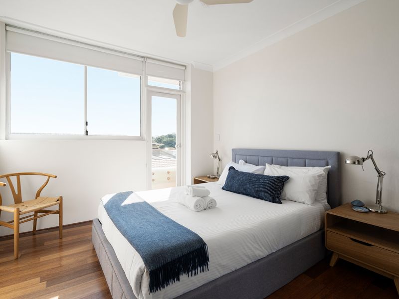 A bright, modern bedroom featuring a grey upholstered bed with blue accents, a wooden nightstand, and a classic wishbone accent chair by the window.