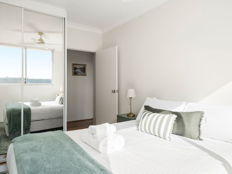Bright bedroom featuring white bedding accented with sage green throws and pillows. A large mirrored sliding closet doors reflect a second bed positioned next to a window, expanding the feel of the compact space.