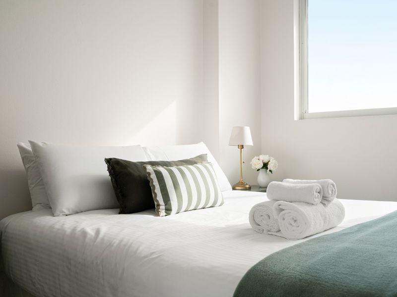 A bright, minimalist bedroom featuring a neatly made bed with white linens, black and white accent pillows, a soft beige headboard, and a simple bedside lamp next to a large window.