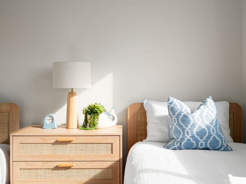 A modern wood-base table lamp sits on the nightstand under soft, natural sunlight.