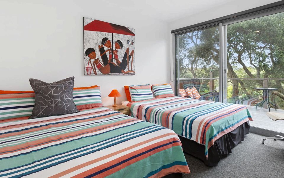 Bedroom 4 featuring two queen beds, a TV, robes, and a sliding door leading on to an outdoor deck equipped with a table and chairs and view of the landscaped back garden.